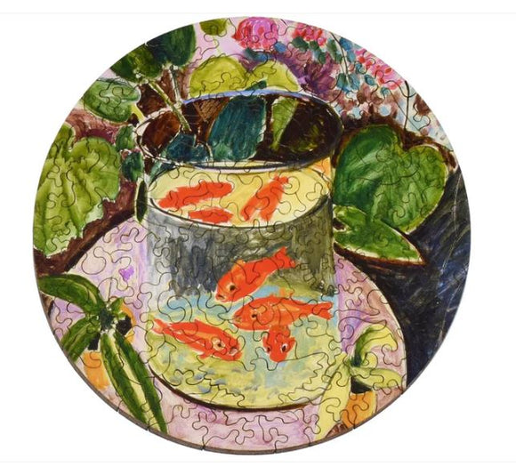 A round jigsaw puzzle depicting a colorful illustration of goldfish swimming in a glass bowl surrounded by green leaves and plants. The art style is reminiscent of Matisse, featuring vibrant hues and fluid lines that create a lively, tranquil scene.
