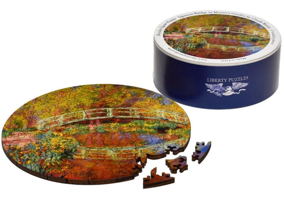 The Japanese Bridge in Monet's Garden jigsaw puzzle by Liberty Puzzles, featuring a vibrant, circular depiction of Monet's iconic bridge surrounded by colorful foliage. The puzzle pieces, displayed partially outside the round puzzle, exhibit intricate designs and colors that echo the original artwork, all housed in a stylish blue circular box.