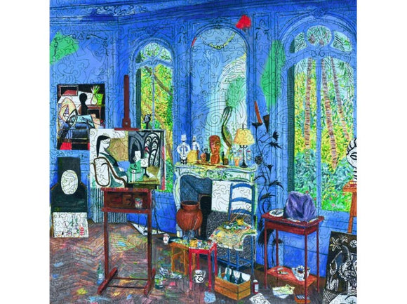A colorful jigsaw puzzle illustration of Picasso's studio, featuring a vibrant blue room filled with art supplies, paintings, and a variety of artistic objects placed throughout the scene. Large windows allow natural light to illuminate the space, while plants are visible outside, creating a creative and lively atmosphere.