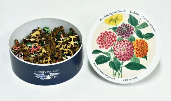 A circular, decorative tin from Liberty Puzzles containing colorful jigsaw puzzle pieces. The lid features an illustration of vibrant dahlias in various colors, showcasing a beautiful floral design. The puzzle is labeled as a wooden jigsaw with 46 pieces, designed for a fun and engaging challenge.