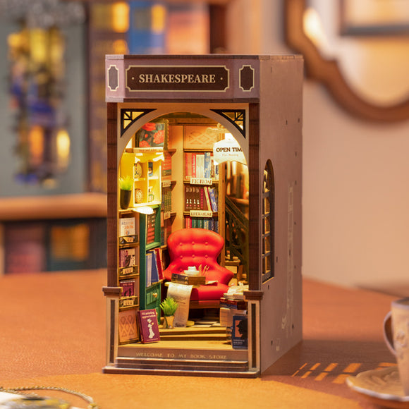A beautifully crafted DIY Book Nook miniature by Robotime, featuring a cozy bookstore setup. The scene includes a bright red armchair, shelves filled with colorful books, and a small reading table. Soft lighting enhances the inviting atmosphere, while a sign indicates 'SHAKESPEARE' above the entrance. Perfect for book lovers and DIY enthusiasts.
