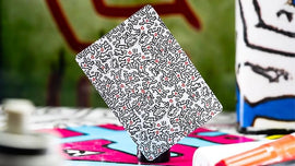 A single playing card featuring a vibrant Keith Haring design, displaying intricate black lines and colorful elements on a white background. The card is showcased in an artistic setting with a blurred background of colorful patterns and art supplies, emphasizing its unique design and appeal for game enthusiasts and art lovers alike.
