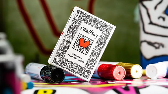 A pack of Keith Haring premium playing cards, decorated with colorful and abstract patterns. The cards are displayed against a vibrant background with art supplies, emphasizing creativity and artistic expression.