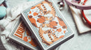 A close-up view of Liquid Cat Playing Cards by 808 Magic and Bacon Playing Co. The deck features whimsical illustrations of playful cats in shades of orange and brown, arranged in a playful design. The cards are stacked on a decorative plate surrounded by colorful cat-themed accessories.