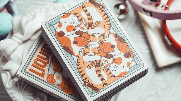 A close-up view of Liquid Cat Playing Cards by 808 Magic and Bacon Playing Co. The deck features whimsical illustrations of playful cats in shades of orange and brown, arranged in a playful design. The cards are stacked on a decorative plate surrounded by colorful cat-themed accessories.