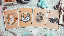 A set of Liquid Cat Playing Cards featuring whimsical illustrations of playful cats, including a cat in a frame, a cat cafe cup, a cat in a fishbowl, and a black cat on a plate. The cards are displayed on a cozy surface with colorful decorative items in the background.