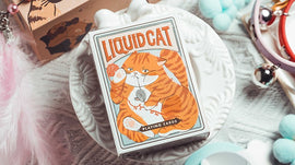 A stylish deck of Liquid Cat Playing Cards by 808 Magic and Bacon Playing Co, featuring a charming and playful illustration of a tabby cat holding a flower. The cards are elegantly designed and presented on a decorative dish surrounded by colorful pom poms and accessories.