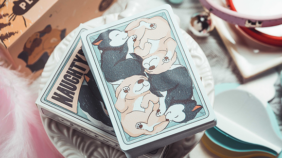 A set of Naughty Dog playing cards designed by 808 Magic and Bacon Playing Co, featuring playful illustrations of various dogs in vibrant colors. The cards are displayed on a decorative plate against a backdrop of game-related items.