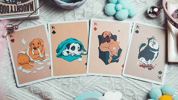 A playful display of Naughty Dog Playing Cards by 808 Magic and Bacon Playing Co. Featuring colorful illustrations of four dog characters on the King cards, showcasing their mischievous antics. The cards are arranged on a textured white fabric surface alongside playful decorative elements, capturing the fun spirit of the game.