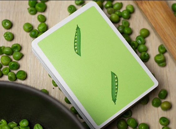 A deck of Sweet Peas Playing Cards by OPC, displayed on a wooden surface surrounded by green peas. The cards feature a vibrant green design with pea pod illustrations, perfect for game nights and family gatherings.