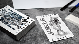 Bicycle Sketch playing cards displayed on a textured surface, featuring a hand-drawn Joker card showing a hand holding a card, alongside a King of Clubs card in an artistic style. The deck is neatly stacked with a textured gray background, pens, and an eraser nearby.