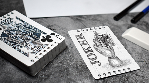 Bicycle Sketch playing cards displayed on a textured surface, featuring a hand-drawn Joker card showing a hand holding a card, alongside a King of Clubs card in an artistic style. The deck is neatly stacked with a textured gray background, pens, and an eraser nearby.