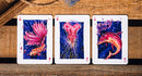 A close-up of three playing cards from the False Anchors V4 (Deep Sea) deck, featuring vibrant illustrations of a goldfish, a jellyfish, and a dolphin against a dark blue background. The cards are laid out on a rustic wooden surface, showcasing their artistic designs and details.