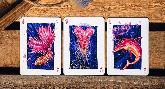 A close-up of three playing cards from the False Anchors V4 (Deep Sea) deck, featuring vibrant illustrations of a goldfish, a jellyfish, and a dolphin against a dark blue background. The cards are laid out on a rustic wooden surface, showcasing their artistic designs and details.