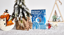 The Snowman Factory Playing Cards by BOCOPO showcased in a festive setting. The box features a whimsical icy blue design with snowman illustrations, surrounded by holiday decorations including a decorative snowman, pine trees, and wooden ornaments.