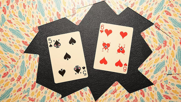 Ace Invaders Playing Cards featuring playful card designs with unique graphics. The set includes illustration of a black spade card (5) and a red heart card (6) amidst colorful patterned backgrounds.
