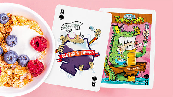 A vibrant display of Frost'd Playing Cards from Howlin' Jack's featuring whimsical designs. The Ace of Spades showcases a quirky character labeled 'Buffed & Puffed,' while another card highlights 'Trapper Snaps,' depicting a colorful scene with cartoon characters enjoying a meal. The cards are set against a pastel pink background with a bowl of cereal topped with blueberries and raspberries.