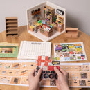 The Fascinating Book Store DIY kit by Robotime showcases a detailed mini bookstore model. The image displays a partially assembled bookstore with bookshelves, a checkout counter, and a variety of books and decorations. There are also assembly instructions and components on the table, including a control panel, emphasizing the engaging and creative nature of this DIY project.