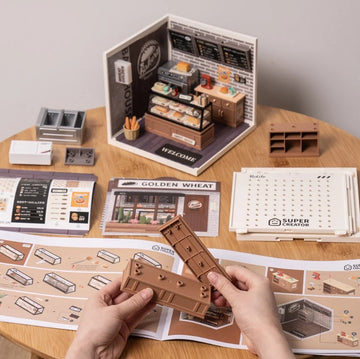 A hands-on DIY kit from Robotime featuring the Golden Wheat Bakery model. The kit includes various components such as miniature furniture, bakery displays, and blueprints laid out on a wooden table, showcasing an engaging crafting experience.