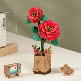 The Red Camelia Model by Robotime is a beautifully crafted DIY kit featuring two vibrant red camellia flowers with green leaves, housed in a decorative pot. The arrangement includes small floral details and a tag that says 'for you', making it a charming decorative item. Nearby, a miniature retro television and a small butterfly figure enhance the scene, adding a playful touch.