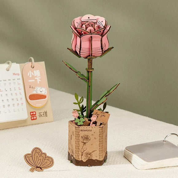 The Pink Rose Model by Robotime is a beautifully crafted DIY kit featuring a detailed pink rose flower atop a green stem, set in a rustic wooden pot. The pot is adorned with small flowers and a tag that says 'for you', providing an elegant touch. Ideal for decoration or as a heartfelt gift, this model combines artistry with a hands-on crafting experience.