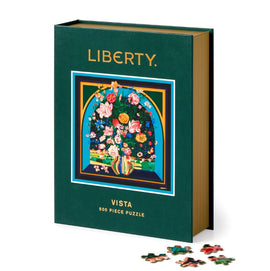 The Liberty Vista jigsaw puzzle by Galison features a vibrant floral design on the box cover, with a colorful arrangement of flowers in a vase, surrounded by a decorative border. The puzzle has 500 pieces and is displayed with some individual pieces scattered in front of the box, showcasing the intricate artwork.