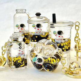 A collection of five astronaut-themed floaty key charms made by BC Mini. Each charm features a small astronaut figure surrounded by golden stars and black liquid, enclosed in transparent containers of various shapes and sizes. The key charms include a bottle, a cup, and other unique designs, highlighting their playful and whimsical nature.