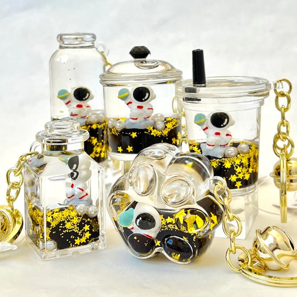 A collection of five astronaut-themed floaty key charms made by BC Mini. Each charm features a small astronaut figure surrounded by golden stars and black liquid, enclosed in transparent containers of various shapes and sizes. The key charms include a bottle, a cup, and other unique designs, highlighting their playful and whimsical nature.