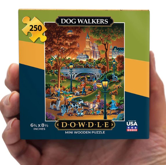 A hand holding the Dowdle Folk Art Mini Dog Walkers jigsaw puzzle box. The box features a colorful illustration of a park scene with various dogs and people, showcasing a vibrant fall backdrop and tall buildings in the distance. The box dimensions are 6.75 x 8.5 inches and includes 250 wooden pieces.
