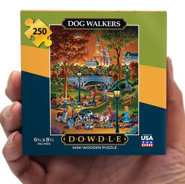 A hand holding the Dowdle Folk Art Mini Dog Walkers jigsaw puzzle box. The box features a colorful illustration of a park scene with various dogs and people, showcasing a vibrant fall backdrop and tall buildings in the distance. The box dimensions are 6.75 x 8.5 inches and includes 250 wooden pieces.