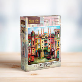 The TBILISI SQUARE jigsaw puzzle by Magnolia Puzzles features a vibrant and colorful illustration of Tbilisi, Georgia, showcasing its unique architecture, including ornate buildings, a clock tower, and lush trees. The packaging highlights the artwork and includes information about the 1023 pieces and its difficulty level. Perfect for puzzle enthusiasts and home decorators.