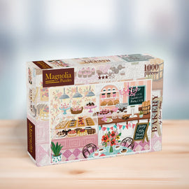 A charming jigsaw puzzle box titled 'BAKERY' by Magnolia Puzzles, featuring a colorful illustration of a bakery display with various desserts, a menu board, and cozy seating. It contains 1000 pieces and showcases intricate details of pastries, cakes, and a floral arrangement.