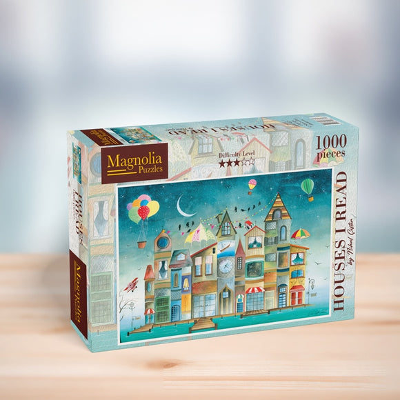 A box of the 'HOUSES I READ' jigsaw puzzle by Magnolia Puzzles. The package features a colorful illustration of whimsical houses with various architectural styles, hot air balloons, and decorations, indicating a fun and engaging puzzle experience. The puzzle contains 1000 pieces and includes a difficulty level rating of 3 stars.