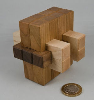Wausau '84 is a wooden mechanical puzzle designed by Jerry Mcfarland. The puzzle features interlocking wooden blocks of various shades and materials, creating a visually striking and challenging design. It is placed next to a coin for size reference, showcasing its intricate craftsmanship and the need for problem-solving skills to disassemble and reassemble the pieces.
