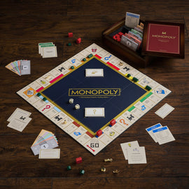 The Monopoly Heritage Edition game set displayed on a wooden table. The board features a deep blue center with the 'MONOPOLY' logo prominently placed. Surrounding the board are various property cards, community chest cards, and money in colorful denominations. Two dice and player tokens are also shown on the board. A neatly arranged wooden box holds additional game cards and pieces, reflecting a classic, nostalgic design. The overall setup invites players to engage in friendly competition.