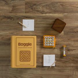 Boggle Vintage Bookshelf Game by Winning Solutions displayed on a wooden surface. The set includes a yellow book-shaped box, letter dice, an hourglass timer, and two notepads with words written on them. Ideal for 2-6 players aged 8 and up.