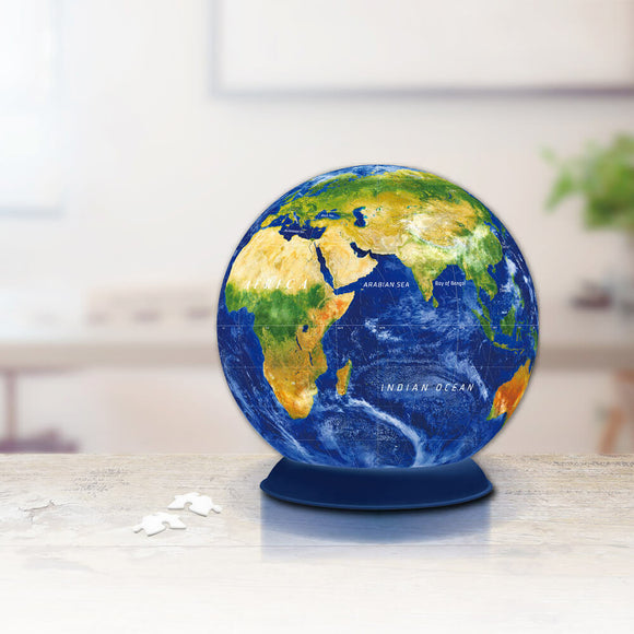 A colorful 3D jigsaw puzzle of Earth, featuring vibrant land and ocean colors. The puzzle is displayed on a blue base with several jigsaw pieces nearby, set against a softly blurred background of a room.