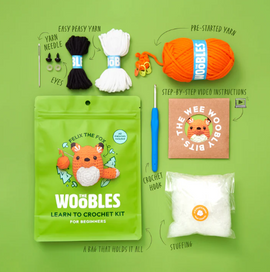 Fox Beginner Crochet Kit from The Woobles, featuring yarn in orange, black, and white, a crochet hook, stuffing, a yarn needle, and step-by-step video instructions. The kit includes a vibrant green packaging with an illustrated fox character, ideal for beginners.