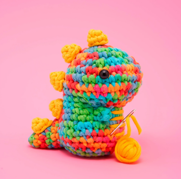 A vibrant crocheted dinosaur in rainbow colors, showcasing bright hues of blue, green, orange, and yellow. It has adorable spines along its back and a cute button eye. A yellow yarn ball and crochet needle are nearby, hinting at a DIY crochet project.