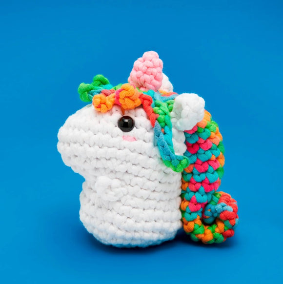 A colorful crocheted unicorn with a white body and vibrant rainbow mane, featuring bright pink, green, and blue yarn. The unicorn has a cheerful expression with black bead eyes and a small pink mouth, sitting against a blue background. Ideal for beginners in crochet, this kit includes all materials needed to create this adorable toy.