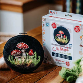 A needle felting kit titled 'Toadstools in A Hoop' by The Crafty Kit Company. The kit features an embroidery hoop containing a colorful felt depiction of mushrooms with red caps and white spots, surrounded by green grass. The product packaging is visible in the background, highlighting the kit's creative potential.