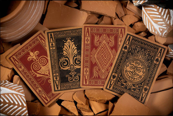 A set of Illiad Playing Cards by Kings Wild displayed on a textured surface with broken terracotta pieces. The cards feature intricate designs in red and black, showcasing artistic patterns and symbols on the Ace cards.
