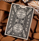 Close-up of the Illiad Playing Cards box by Kings Wild, featuring intricate black and white designs with circular graphics depicting creative faces and floral patterns, set against a background of textured brown surfaces.