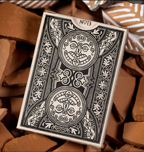Close-up of the Illiad Playing Cards box by Kings Wild, featuring intricate black and white designs with circular graphics depicting creative faces and floral patterns, set against a background of textured brown surfaces.