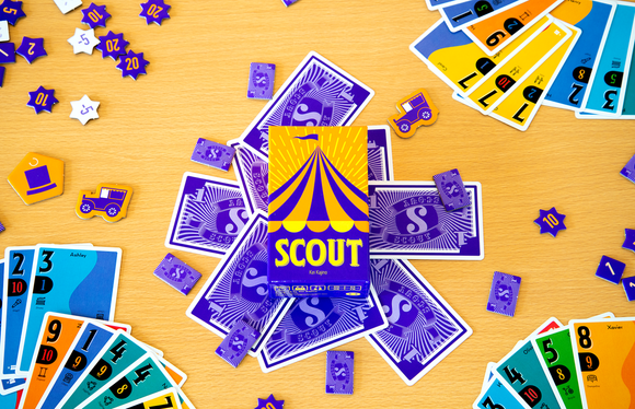 The game Scout by OINK GAMES features a vibrant box prominently displayed in the center, surrounded by colorful cards, game tokens, and play money. The cards showcase numbers and various designs, while the tokens are star-shaped and feature different values. The game is laid out on a wooden surface, creating an inviting atmosphere for gameplay.