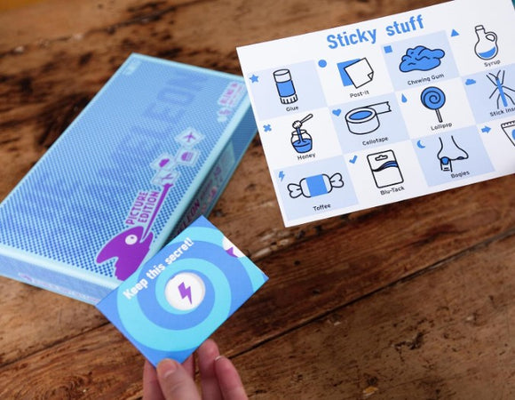 The Chameleon: Pictures Edition game by Big Potato Games is displayed on a wooden table. The blue box features colorful graphics, and a hand is holding a blue card that says 'Keep this secret!' Next to it is a card listing various 'sticky stuff' like glue, honey, and tape with corresponding icons.