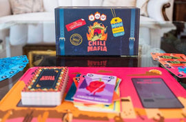 Chili Mafia Deluxe Edition game box displayed on a table. The game features colorful cards, including a red chili card, and a vibrant game board. The box is decorated with bold graphics and text promoting the game's spicy theme. Nearby accessories include player boards and a rulebook, creating an inviting game setup for friends and family.