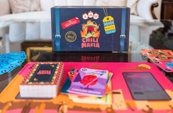 Chili Mafia Deluxe Edition game box displayed on a table. The game features colorful cards, including a red chili card, and a vibrant game board. The box is decorated with bold graphics and text promoting the game's spicy theme. Nearby accessories include player boards and a rulebook, creating an inviting game setup for friends and family.