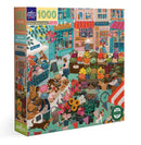The English Green Market 1000-piece jigsaw puzzle by Eeboo features a vibrant and colorful illustration of a bustling market scene. Rich in detail, the artwork depicts various stalls filled with fresh fruits, flowers, and baked goods. People are seen enjoying the market, with pets and bicycles adding charm to the lively atmosphere. The puzzle box emphasizes its eco-friendly materials.
