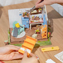 A DIY kit showcasing the Dreamy Garden House by Robotime. The image features a miniature interior with vibrant furniture, including a blue cabinet, a cozy green chair, and a rustic table set up with various decorations and plants. Hands are seen assembling a colorful orange shelf, highlighting the intricate details and craftsmanship of the model.